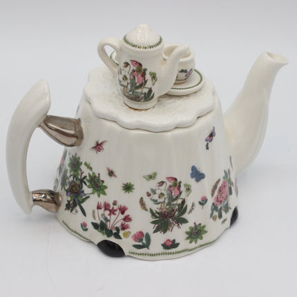 A vintage white porcelain teapot decorated with colorful floral and butterfly motifs, featuring a matching small teapot and cup on top, photographed on a white background.