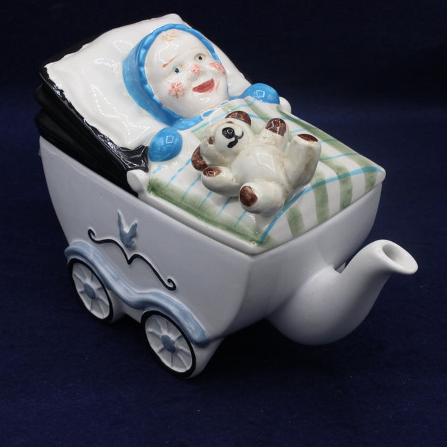 Ceramic teapot shaped like a baby carriage with a baby and dog figure on a dark background