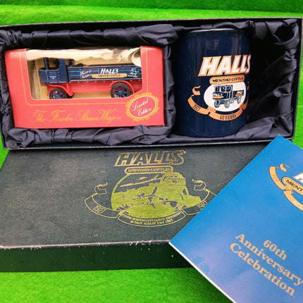 A boxed Halls Midland Railway 60th Anniversary gift set on a bright green background. The set includes a miniature model train lorry in a clear window box, a dark blue ceramic mug with Halls branding, a black presentation box with gold crest, and a blue booklet.