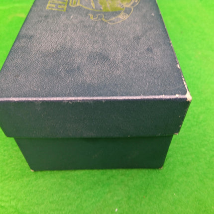 A dark textured shoe box with a gold logo on top, photographed on a bright green background.