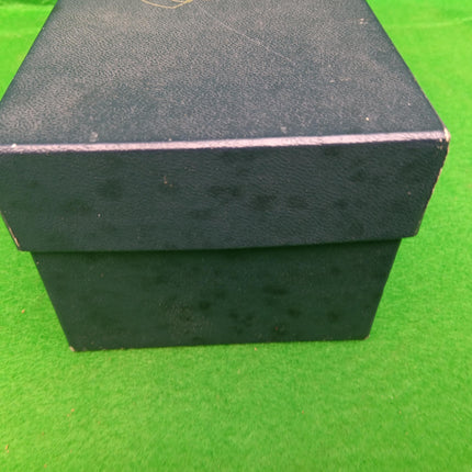 A black textured gift box with a hinged lid, photographed on a bright green artificial turf background.
