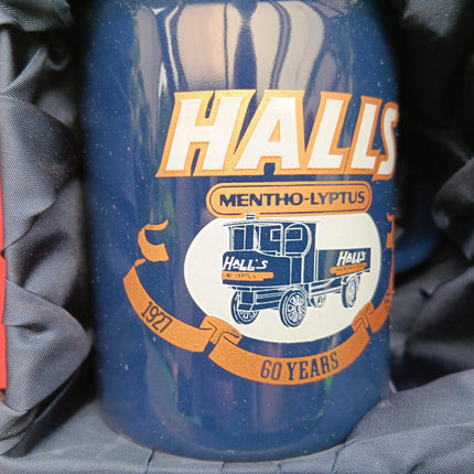 A blue ceramic mug with a vintage-style Halls Mentho-Lyptus label, nestled in dark gray fabric packaging. The label features white and orange lettering and a retro truck illustration celebrating 60 years.