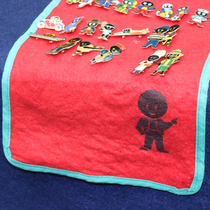 A red fabric display cloth with a turquoise border, featuring a black silhouette of a Golly on the lower right and several colorful Golly enamel pin characters arranged along the top. The cloth is laid on a dark blue surface.