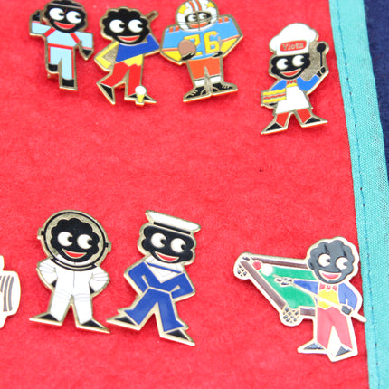 A close-up of several colorful enamel Robertson's Golly character pins arranged on a bright red fabric surface. The Golly pins feature cartoon-style figures in various outfits, including sports, space, and traditional clothing, with gold-tone metal outlines.
