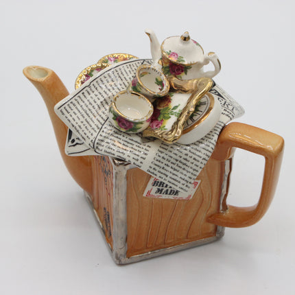 A novelty ceramic teapot shaped like a wooden chest, with a miniature floral teapot, teacups, saucer, and gold spoon resting on top of printed newspaper detailing. The teapot features a warm brown wood-grain finish with silver-painted edges and a small 'Handmade' label.