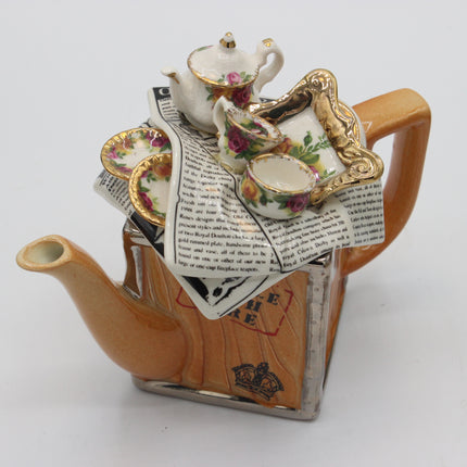 A decorative teapot-shaped ceramic object with a peach-brown body, silver-toned base, and a miniature tea set arrangement on top featuring white teapot and cups with pink floral detailing and gold trim, set against a plain light background.