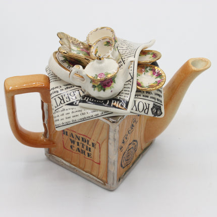 A decorative ceramic teapot with a warm orange-brown body and spout, featuring a printed newspaper design on the front. The top is decorated with miniature white teacups and saucers, some printed with floral motifs, resting on top of folded newspaper pieces.