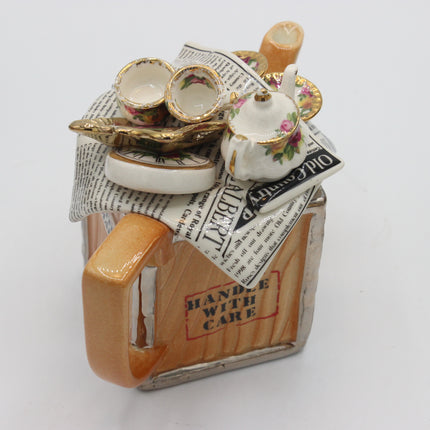 A vintage-style ceramic teapot figurine with a wooden-textured body, orange-brown handle and base, and a floral teacup set on top with newspaper accents.