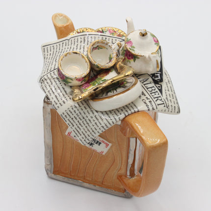 A ceramic novelty teapot shaped like a beer mug, decorated with a floral patterned tea set arrangement on top, including teacups, a teapot, a gold spoon, and a newspaper print fabric draped over the opening, photographed on a white background.