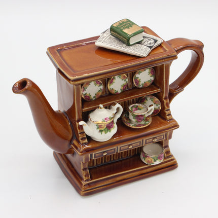 A brown ceramic teapot shaped like a miniature tea cabinet, with floral teacups on the shelves, a small white teapot, a book, and a newspaper on top, photographed on a white background.