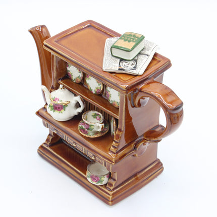 A miniature ceramic tea set display shaped like a brown wooden tea cart, featuring a white teapot, teacups, saucers, and a small book on top, photographed on a white background.