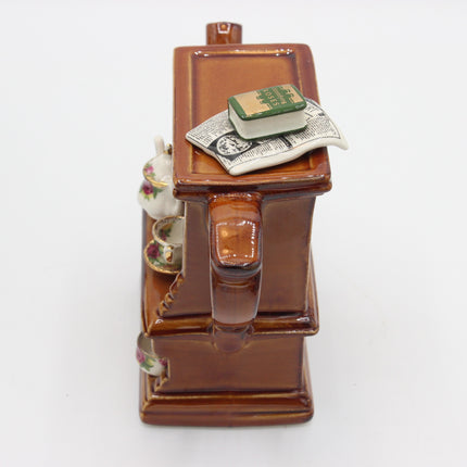 A brown glazed ceramic teapot shaped like a miniature bookshelf or cabinet, with a small book and dollar bills on top, photographed on a plain white background.