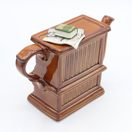 A brown ceramic novelty pitcher shaped like a miniature cabinet or book, with a handle on the left and a small spout on top. A folded dollar bill and a miniature green book rest on the lid.