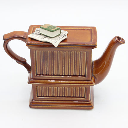 A brown ceramic novelty teapot shaped like a book, featuring a ribbed cover, a curved handle, a spout, and a small miniature book and dollar bill decoration on top, set against a plain white background.