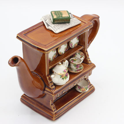 A brown ceramic miniature tea set shaped like a cabinet, featuring a teapot, teacups, saucers, and a small box on top, photographed on a white background.