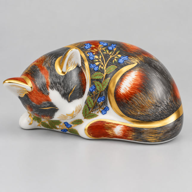 A decorative porcelain figurine of a sleeping cat, decorated with realistic fur patterns in black, red, and white, accented with blue flowers, green leaves, and gold metallic trim, set against a plain light gray background.
