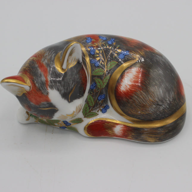 A decorative porcelain figurine of a sleeping cat, decorated with realistic fur patterns in black, red, and white, accented with blue flowers, green leaves, and gold metallic trim, set against a plain light gray background.