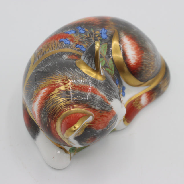 A hand-painted porcelain cat figurine curled up in a sleeping pose, decorated with red, white, black, blue, and green floral details and gold accents, on a plain white background.