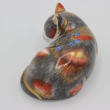 A decorative cat figurine or small sculpture shown on a plain light gray background. The cat is curled up and painted with a mottled gray, black, and white fur pattern, accented with red patches, blue flowers, and gold outlines.