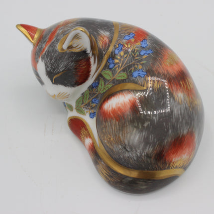 A ceramic cat figurine or box shaped like a curled sleeping cat, decorated with realistic fur patterns in black, gray, red, and white, blue flowers, green leaves, and gold trim, photographed on a plain light background.