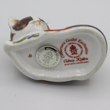 A vintage Royal Crown Derby Camip Kitten ceramic figurine shown from its underside, featuring a white glazed body with a multicolored kitten head, red collector's guild inscription, and a silver coin insert.