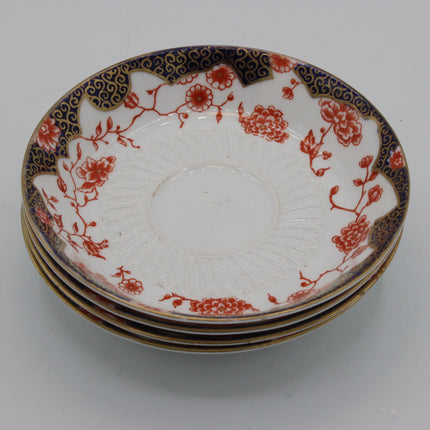 A stack of four vintage crown derby ceramic plates with a white glaze, decorated with red floral patterns and a dark blue border with gold detailing, set against a plain light gray background.