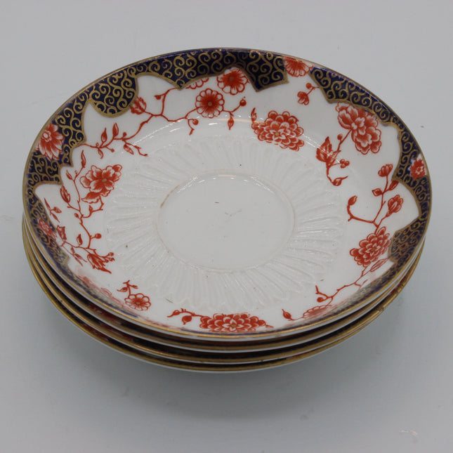 A stack of four vintage crown derby ceramic plates with a white glaze, decorated with red floral patterns and a dark blue border with gold detailing, set against a plain light gray background.