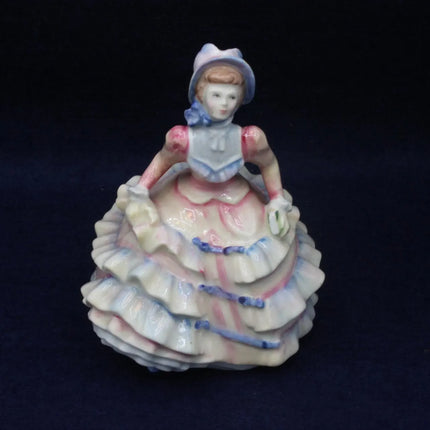 A vintage porcelain figurine of a seated young woman in a ruffled white dress with pink and blue accents, set against a dark navy background.