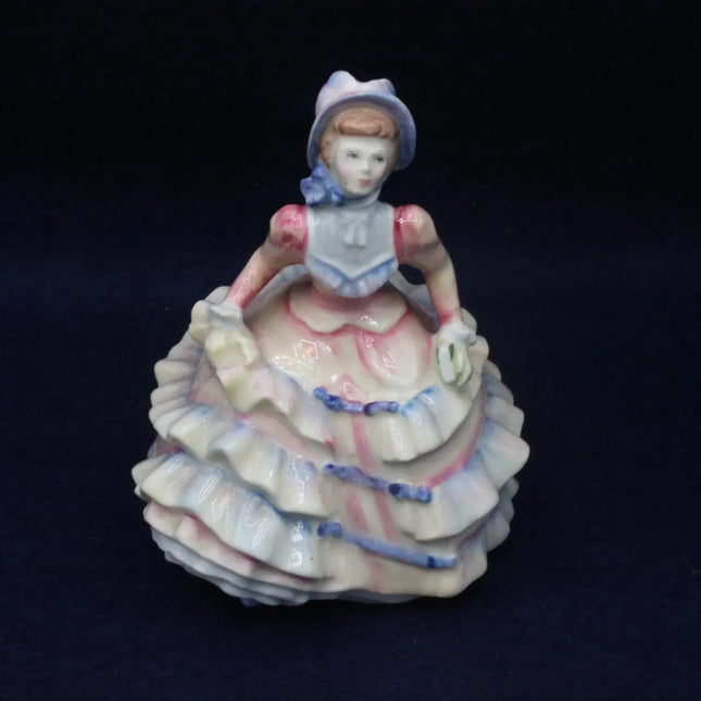 A vintage porcelain figurine of a seated young woman in a ruffled white dress with pink and blue accents, set against a dark navy background.