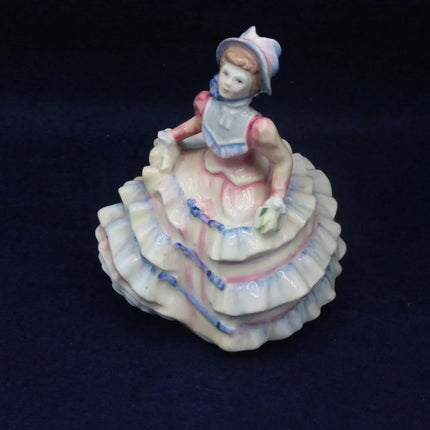 A vintage porcelain figurine of a young girl in a tiered dress, displayed on a dark navy blue background.