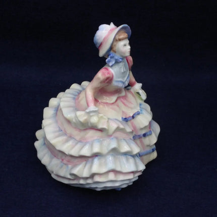 A vintage porcelain figurine of a young girl in a ruffled white dress with pink and blue accents, set against a dark navy background.