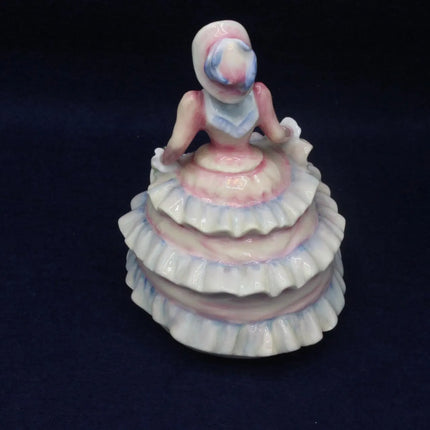 A vintage ceramic figurine of a ballerina with a pink and blue painted face, wearing a tiered white, pink, and blue dress, set against a dark navy blue background.