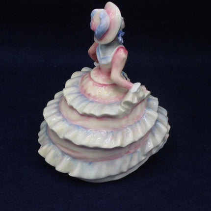 A vintage porcelain figurine of a seated woman wearing a tiered ruffled dress and hat, decorated with pink, blue, and white glazes, set against a dark navy background.
