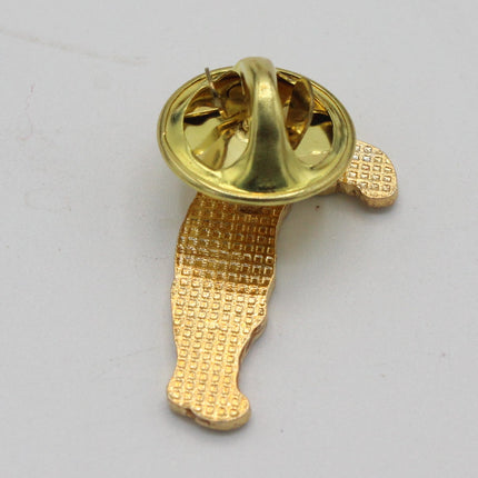 A gold-colored enamel pin showing its back side with the clasp mechanism, placed on a plain light gray background.