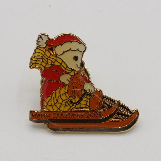 A small enamel lapel pin showing a teddy bear in a red Santa hat and red coat riding a sled, with the text 'Merry Christmas 2003' on the front, photographed on a plain light background.
