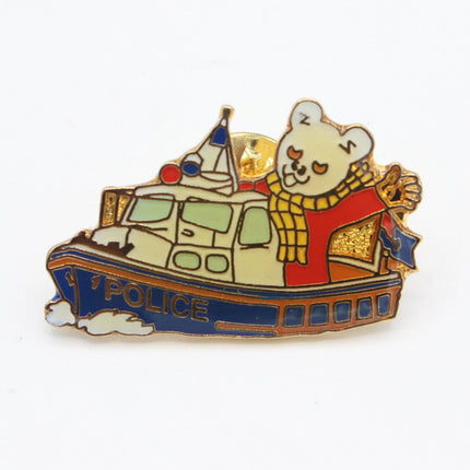 A small enamel pin shaped like a police boat, featuring a cream-colored bear character wearing a red sweater and yellow scarf, with the word 'POLICE' on the side of the blue boat, shown on a plain white background.