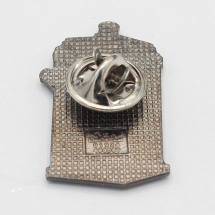 A small silver-toned enamel pin shaped like a house, shown from the back on a plain white background. The pin features a textured patterned face and a circular pin clasp on top.