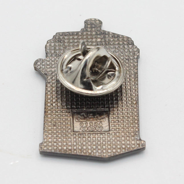 A small silver-toned enamel pin shaped like a house, shown from the back on a plain white background. The pin features a textured patterned face and a circular pin clasp on top.