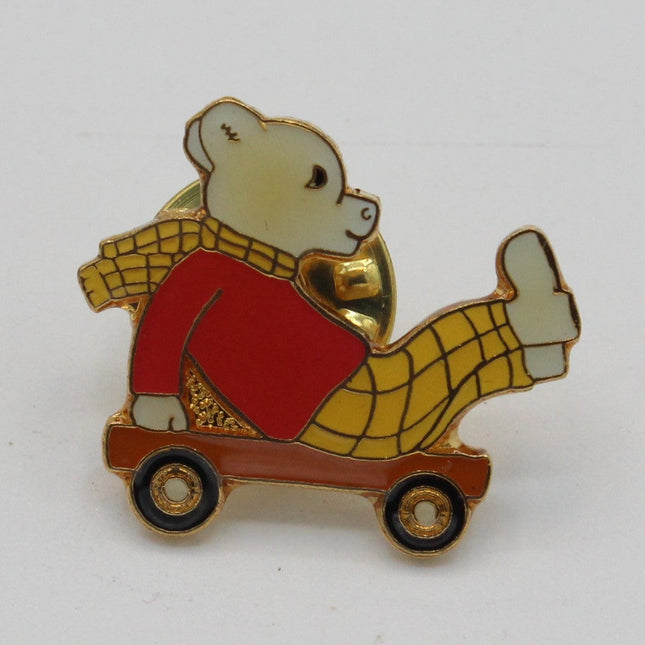 A small enamel pin depicting a teddy bear wearing a red sweater and yellow plaid pants, sitting on a brown toy cart, set against a plain white background.