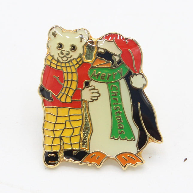 A small enamel pin featuring a teddy bear in a red sweater and yellow plaid pants hugging a penguin wearing a red hat and green scarf, on a plain white background.