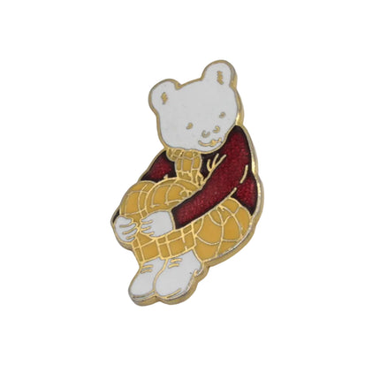 A small enamel pin shaped like Rupert the  bear wearing a yellow checkered outfit and a dark red sweater, shown on a plain light gray background.