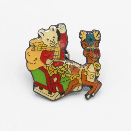 A small enamel pin depicting Rupert Bear as Santa Claus in a red coat and yellow scarf riding a brown reindeer on a sleigh, shown on a plain white background.