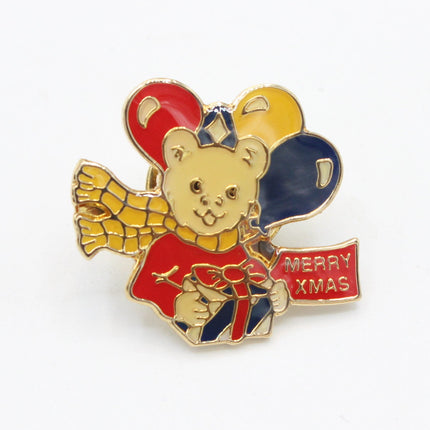 A small enamel pin shaped like a teddy bear holding red, yellow, and blue balloons, with a red banner that reads 'MERRY KMAS', photographed on a plain white background.
