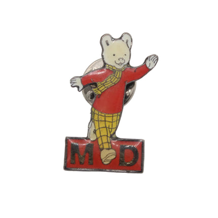 A vintage-style enamel pin featuring a Rupert bear wearing a red sweater and yellow plaid trousers, standing on a red block with the letters 'M D' in black, photographed against a plain white background.