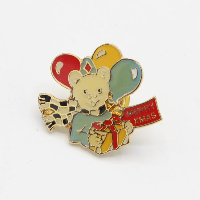 A small enamel pin featuring a teddy bear holding balloons, wrapped in a scarf, and holding a red banner that says 'HAPPY XMAS', set against a plain white background.