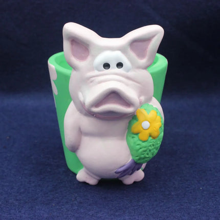 A small pig figurine standing in front of a green cylindrical planter, holding a yellow flower. The pig is pale pink with purple feet and ears, and the planter is green with white spots.