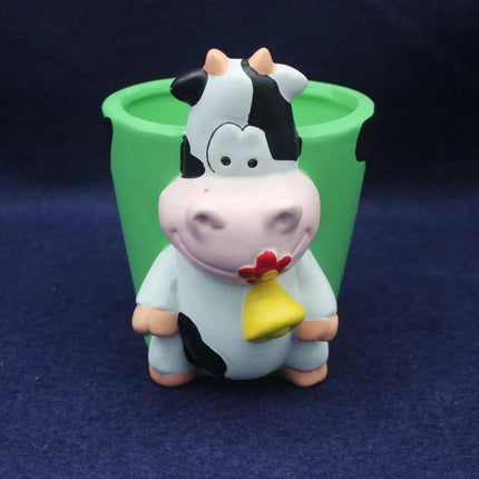 A small cow figurine with a black-and-white pattern, pink face, and yellow bell, standing in front of a green cup on a dark blue background.