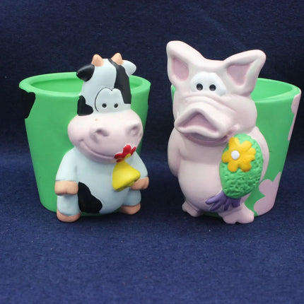 Two small novelty figurines shaped like a black-and-white cow and a pink pig, each standing in front of a green cup-like container, photographed on a dark blue fabric background.