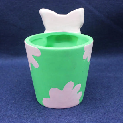 A green ceramic cup with a white cat-shaped lid and white cloud-like patterns, set against a dark blue textured background.