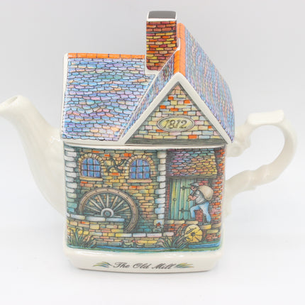 A decorative ceramic teapot shaped like a vintage watermill, featuring a colorful illustrated facade with a water wheel, windows, a chimney, and the text 'The Old Mill' on the base, photographed against a plain light gray background.